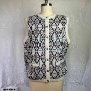 Könnte beinhalten: Eine ärmellose Strickweste mit Knöpfen, cremefarben und mit einem geometrischen Muster in Marineblau, Rot und Weiß. Die Weste hat einen Rundhalsausschnitt, zwei Vordertaschen und dunkelgraue Knöpfe. Die Weste wird auf einer Schaufensterpuppe präsentiert.