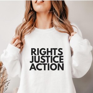 Könnte beinhalten: Weißes Sweatshirt mit dem Aufdruck "RIGHTS JUSTICE ACTION" in fetten schwarzen Buchstaben. Das Sweatshirt hat einen Rundhalsausschnitt und scheint aus einem weichen Material zu sein. Der Gesamteindruck ist lässig und bequem.