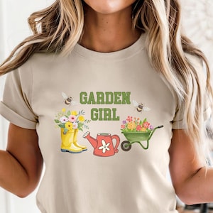 Könnte beinhalten: Beiges T-Shirt mit dem Text "GARDEN GIRL" in Grün. Das Shirt zeigt Illustrationen eines gelben Gummistiefels mit Blumen, einer roten Gießkanne, einer grünen Schubkarre und zwei Bienen.