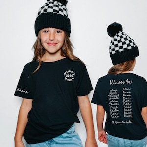 Op de afbeelding: Een zwart t-shirt met een witte cirkelvormige afbeelding op de borst met de tekst GRUNDSCHULE 2026 GESCHAFFT. Op de mouw staat de naam Emelie. De achterkant toont de tekst Klasse 4a, een lijst met namen en 18. Grundschule.