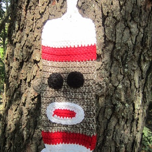 May include: A crocheted monkey-face bottle holder with a white and red striped design. The holder has a brown face with black eyes and a red mouth.