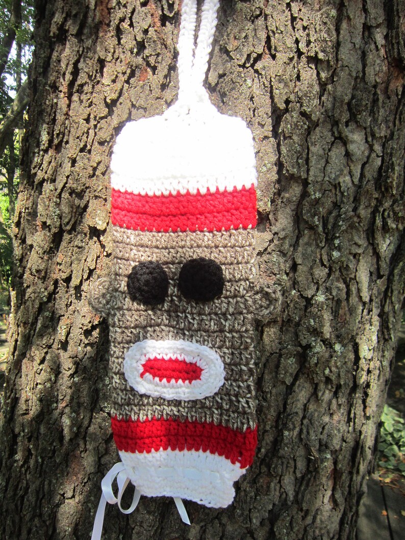 May include: A crocheted monkey-face bottle holder with a white and red striped design. The holder has a brown face with black eyes and a red mouth.