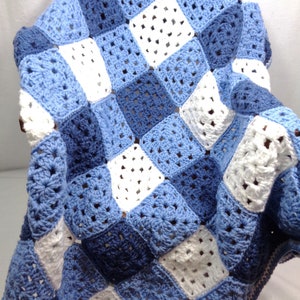 May include: A handmade crochet blanket featuring a patchwork design. The blanket is composed of squares in shades of blue and white. The squares are arranged in a grid pattern, creating a visually appealing and textured surface.
