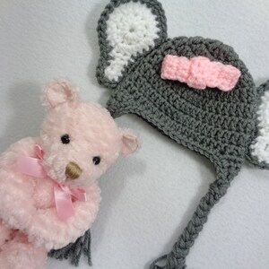 May include: A gray crocheted elephant hat with white ears and a pink bow. The hat has a long tail that can be tied under the chin. The hat is sitting next to a pink teddy bear.