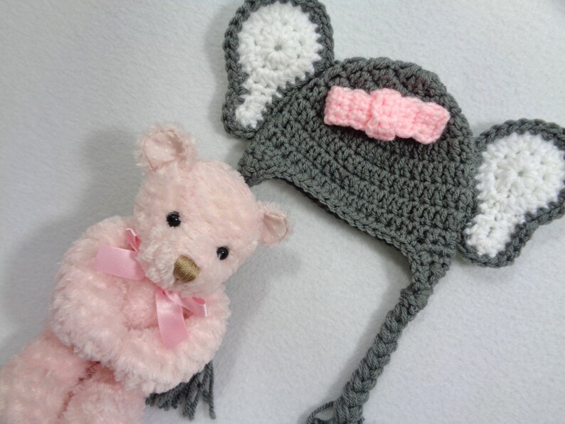 May include: A gray crocheted elephant hat with white ears and a pink bow. The hat has a long tail that can be tied under the chin. The hat is sitting next to a pink teddy bear.
