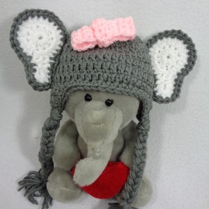 May include: A gray crocheted elephant hat with white ears and a pink bow. The hat has long gray braids that hang down the sides. The hat is being worn by a gray plush elephant toy holding a red heart.
