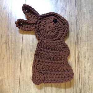 May include: A handmade brown crochet bunny with a round body, two long ears, and a small black eye. The bunny is lying on a wooden surface. This item is perfect for Easter decorations or as a child's toy.