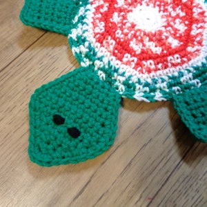 Pode incluir: Um descanso de panela de tartaruga de crochê com um esquema de cores vermelho, branco e verde. A tartaruga tem uma carapaça verde com um centro branco e vermelho e pernas verdes.