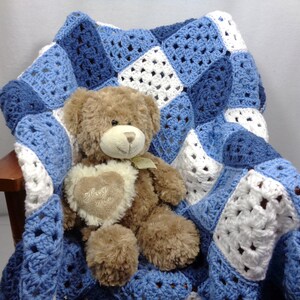 May include: A handmade crochet blanket in shades of blue and white, draped over a wooden chair. A brown teddy bear with a heart-shaped patch that says "Hug Me" is nestled on the blanket. The blanket features a granny square design.
