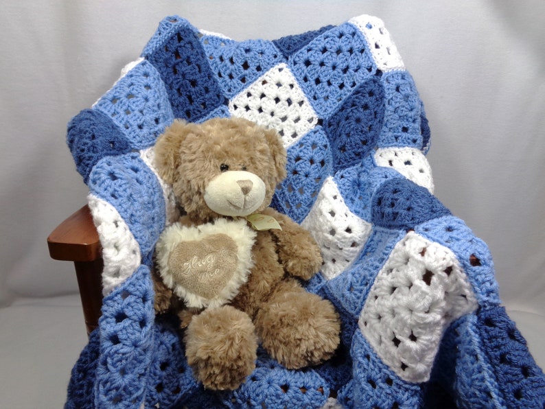 May include: A handmade crochet blanket in shades of blue and white, draped over a wooden chair. A brown teddy bear with a heart-shaped patch that says "Hug Me" is nestled on the blanket. The blanket features a granny square design.