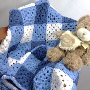 May include: A handmade crochet blanket in shades of blue and white, draped over a wooden chair. The blanket features a granny square design. A brown teddy bear with a cream-colored patch rests on the blanket.