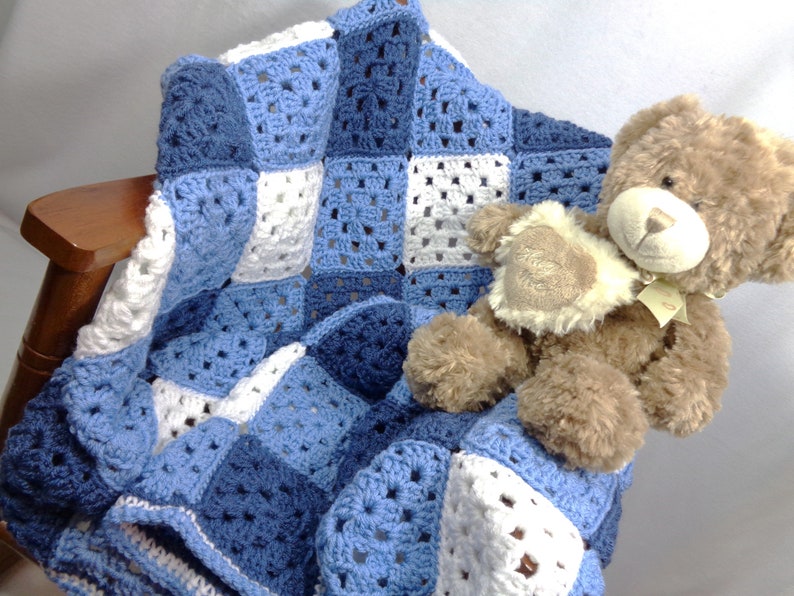 May include: A handmade crochet blanket in shades of blue and white, draped over a wooden chair. The blanket features a granny square design. A brown teddy bear with a cream-colored patch rests on the blanket.