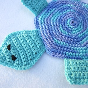 Puede incluir: Un juguete de tortuga de crochet con una concha azul y morada y extremidades verde turquesa. La tortuga tiene ojos negros.