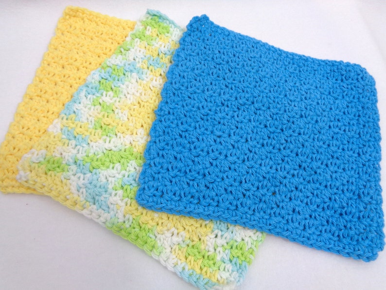May include: Three crocheted dishcloths in yellow, green, white, and blue. The dishcloths are made with a simple crochet stitch.