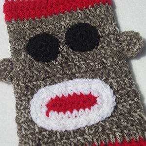 May include: Crocheted monkey sock puppet with a red, white, and brown color scheme. The puppet has a large, open mouth with white teeth and a red tongue.