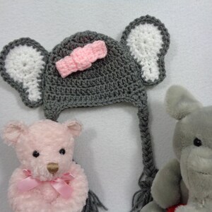 May include: A gray crocheted elephant hat with white ears and a pink bow. The hat has a long tail that can be tied under the chin. The hat is sitting on a white surface next to two stuffed animals.