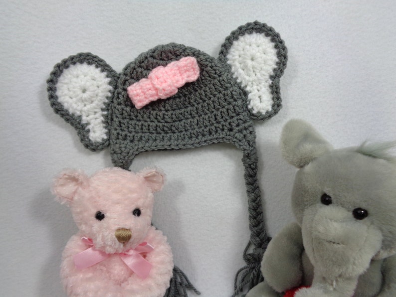 May include: A gray crocheted elephant hat with white ears and a pink bow. The hat has a long tail that can be tied under the chin. The hat is sitting on a white surface next to two stuffed animals.