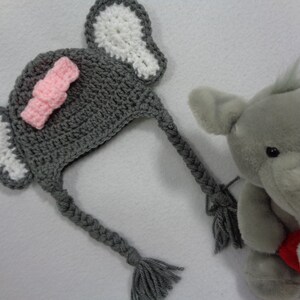 May include: Gray crocheted elephant hat with pink bow and white ears. The hat has a long gray braid that can be tied under the chin.