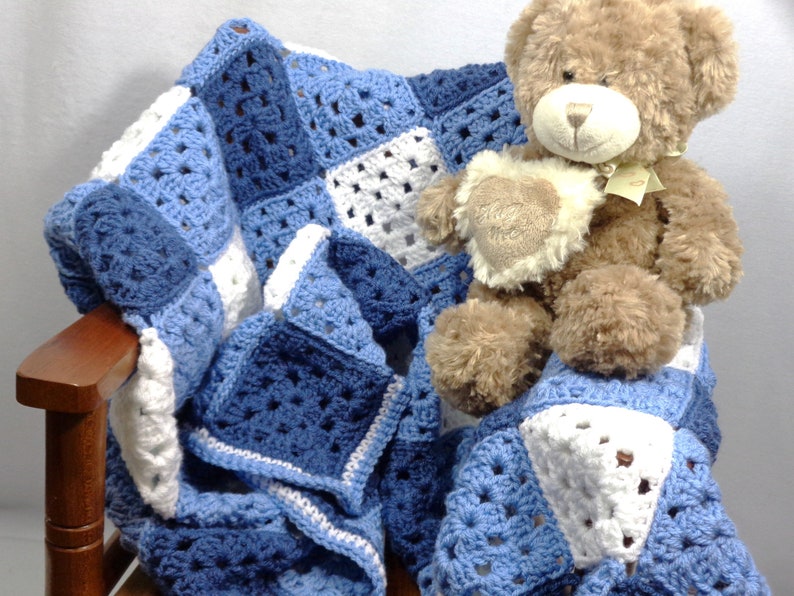 May include: A handmade crochet blanket in shades of blue and white, draped over a wooden chair. A plush teddy bear with a heart-shaped accent sits atop the blanket. The blanket features a square pattern design, perfect for a baby or child.