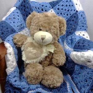 May include: A brown teddy bear with a cream-colored heart-shaped patch that says "Hug Me" is nestled on a blue, white, and light blue crocheted blanket. The blanket is draped over a wooden chair.