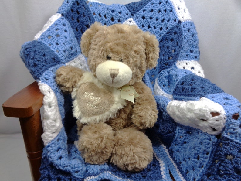 May include: A brown teddy bear with a cream-colored heart-shaped patch that says "Hug Me" is nestled on a blue, white, and light blue crocheted blanket. The blanket is draped over a wooden chair.