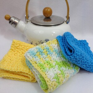 May include: Three crocheted dishcloths in yellow, green, white, and blue. The dishcloths are arranged in front of a white enamel teapot with a wooden handle and knob.