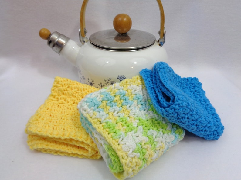 May include: Three crocheted dishcloths in yellow, green, white, and blue. The dishcloths are arranged in front of a white enamel teapot with a wooden handle and knob.