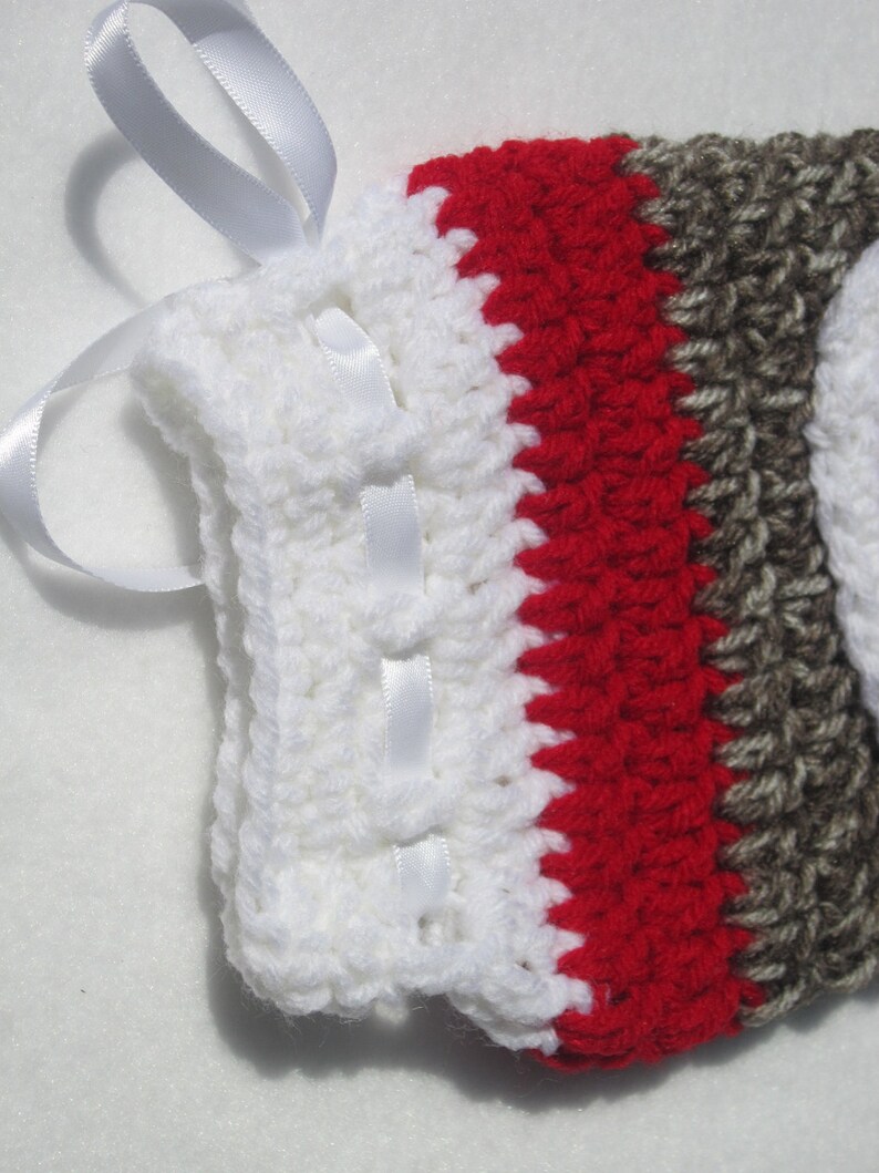 May include: White crocheted boot cuff with red and brown stripes and a white satin ribbon tied around the top.
