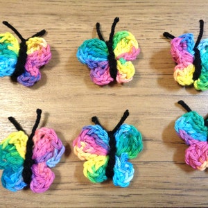 Könnte beinhalten: Sechs gehäkelte Schmetterlinge in Regenbogenfarben mit schwarzen Garnantennen. Die Schmetterlinge sind in zwei Reihen zu je drei auf einer Holzoberfläche angeordnet.