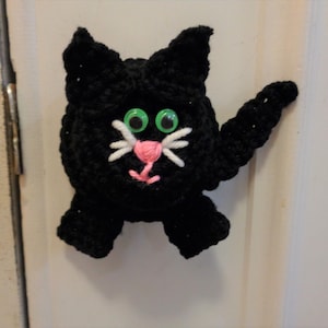 Może przedstawiać: Czarny, szydełkowy kot z zielonymi oczami i różowym nosem. Kot trzyma prawą łapę w górze.