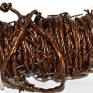 Op de afbeelding: Een spoel bruin prikkeldraad. De draad is strak opgerold en heeft scherpe weerhaken. De draad heeft een metallic bruine kleur en zit op een transparante plastic spoel. De draad wordt waarschijnlijk gebruikt voor afrastering of decoratieve doeleinden.