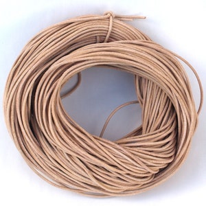 Puede incluir: Un rollo de cordón de cuero de color marrón claro natural. El cordón es fino y redondo, con una superficie ligeramente texturizada. El rollo está atado en un extremo con un trozo más pequeño del mismo cordón.