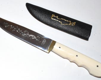 Puede incluir: Un cuchillo de acero inoxidable con mango color crema y funda negra. La hoja del cuchillo tiene un diseño floral grabado. El mango tiene forma curva con tres puntos negros. La funda tiene un elemento decorativo dorado.