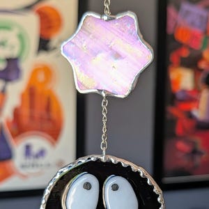 May include: A stained glass suncatcher featuring a black, round character with large white eyes and a star-shaped top piece. The star and the character are connected by a silver chain. The suncatcher is designed to hang and catch the light.
