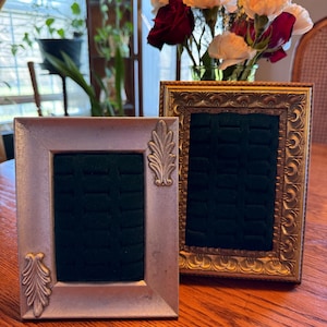 May include: Two decorative picture frames with ornate detailing. One frame is silver with leaf accents, the other is gold with a detailed border. Both frames have a dark green velvet interior, likely for displaying jewelry or small items.