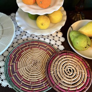 May include: Two round woven placemats with concentric circles in shades of tan, pink, and green. The placemats are on a dark surface, with a white tiered serving dish filled with oranges, lemons, and limes in the background.