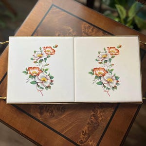 May include: A gold metal serving tray with two white ceramic tiles featuring a floral design of white and orange roses. The tiles are framed with a gold trim.