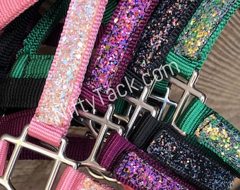May include: A collection of nylon horse headcollars featuring silver-toned metal hardware and glittery overlays in pink, purple, black, and green. The text "glittytack.com" is printed across the centre of the overlapping straps in white lettering.