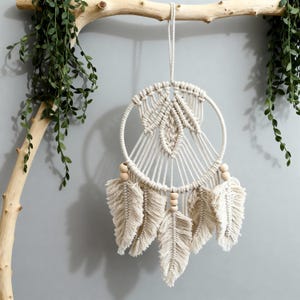 Peut inclure: Attrape-rêves en macramé crème avec un cadre circulaire et un centre tissé. Il comporte trois accents de plumes suspendues et des perles en bois. L'attrape-rêves est suspendu à une branche naturelle, avec un feuillage vert en arrière-plan.