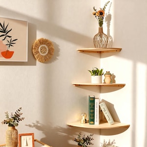 Könnte beinhalten: Drei hölzerne Eckregale an einer weißen Wand, die jeweils dekorative Gegenstände präsentieren. Das obere Regal zeigt eine Vase mit Blumen, das mittlere Regal hat eine Topfpflanze und eine Froschfigur, und das untere Regal enthält Bücher und eine kleine Lampe.