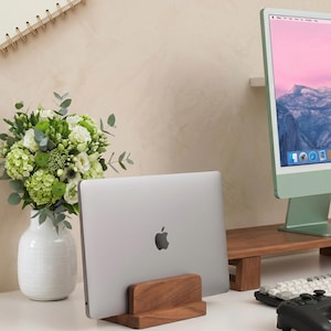 Peut inclure: Un ordinateur portable argenté avec un logo Apple noir est placé dans un support en bois. Un bouquet de fleurs blanches et vertes est dans un vase blanc. Un écran d'ordinateur vert et un clavier sont également visibles.