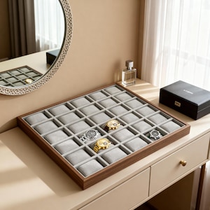 May include: A wooden watch display case with a gray interior and individual compartments. Several watches with gold and silver bands are displayed. The case sits on a light-colored dresser with a mirror and perfume bottle in the background.