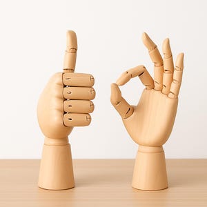 Könnte beinhalten: Zwei hölzerne, bewegliche Hände, eine mit erhobenem Daumen und die andere mit dem „Okay“-Zeichen. Die Hände sind hellbraun und stehen auf einer hellen Holzoberfläche vor einem weißen Hintergrund. Die Hände sind für künstlerische oder dekorative Zwecke bestimmt.