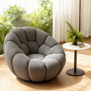 May include: A grey, textured, round accent chair with a unique, segmented design. The chair sits on a black metal base. A small, white side table with a fern and a book is next to the chair.