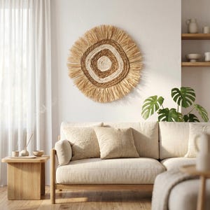 May include: A circular woven wall hanging with a fringed edge, in natural beige and cream tones. The wall art features concentric circles, creating a textured, bohemian style. It hangs above a beige sofa with matching cushions, complementing the room's neutral colour scheme.