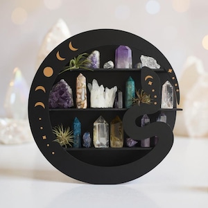 Peut inclure: Étagère noire circulaire en forme de serpent, contenant divers cristaux et petites plantes. L'étagère comporte trois niveaux et est ornée de croissants de lune et d'étoiles dorés. Les cristaux sont de couleurs violet, transparent, bleu et jaune.