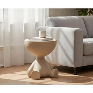 Pode incluir: Uma mesa lateral bege com uma forma única semelhante à humana. A mesa tem um tampo arredondado com um livro e uma caneca. A base da mesa tem a forma de uma figura sentada. A mesa está colocada ao lado de um sofá cinza em uma sala bem iluminada.