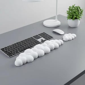 Peut inclure: Un repose-poignet et un tapis de souris ergonomiques blancs en forme de nuages sont posés sur un bureau gris, à côté d'un clavier et d'une souris. Une petite plante en pot et une lampe de bureau blanche sont également visibles.