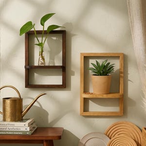 Peut inclure: Deux étagères murales en bois, une marron foncé et une marron clair, chacune avec une petite étagère. L'étagère marron foncé contient un vase en verre avec des plantes vertes. L'étagère marron clair contient une plante en pot. Un arrosoir doré et des livres sont sur une table.
