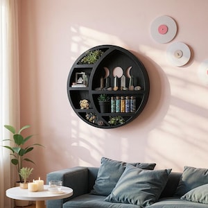 Puede incluir: Estantería de pared circular negra con diseño de fases lunares, llena de plantas, cristales y objetos decorativos. La estantería está montada en una pared rosa claro. La imagen también muestra un sofá y una mesa auxiliar.